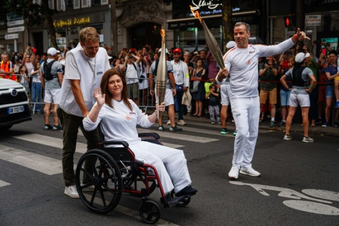 Lebanese photojournalist Christina Assi carries the Olympic torch, to honor journalists wounded and killed in the field, in Vincennes, near Paris, France, on July 21, 2024. Christina Assi, of Agence France-Presse or AFP, was among six journalists struck by Israeli shelling on October 13th, 2023 while reporting on Southern Lebanon, Reuters photographer Issam Abdallah was killed in the attack. AFP videographer Dylan Collins, also wounded in the same attack, is pushing Assi's wheelchair. Photo by Ammar Abd Rabbo/ABACAPRESS.COM